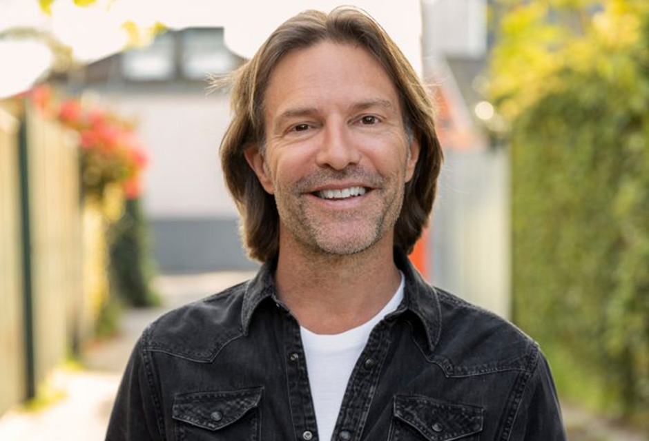 A headshot of Eric Whitacre, a white man with light brown shoulder length hair, wearing a denim jacket, smiling at the camera.